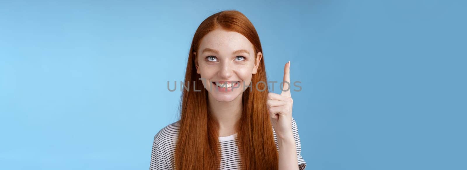 Intrigued lively happy smiling redhead european girl 20s look pointing up index fingers amused check out interesting sale holiday promo offer standing intrigued thrilled blue background. Lifestyle.