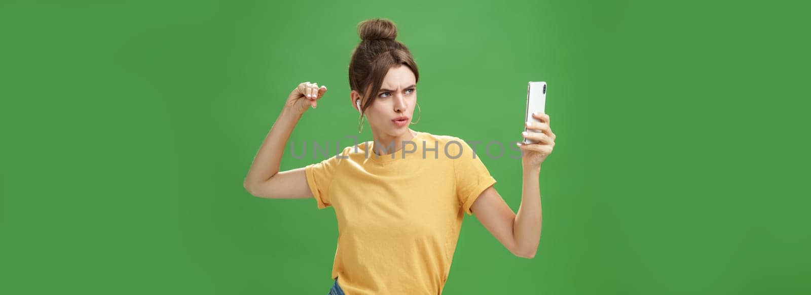 Woman making serious look to take selfie while listening music in wireless earphones posing and staring at smartphone screen daring and aggressive, liking post photos online over green background by Benzoix