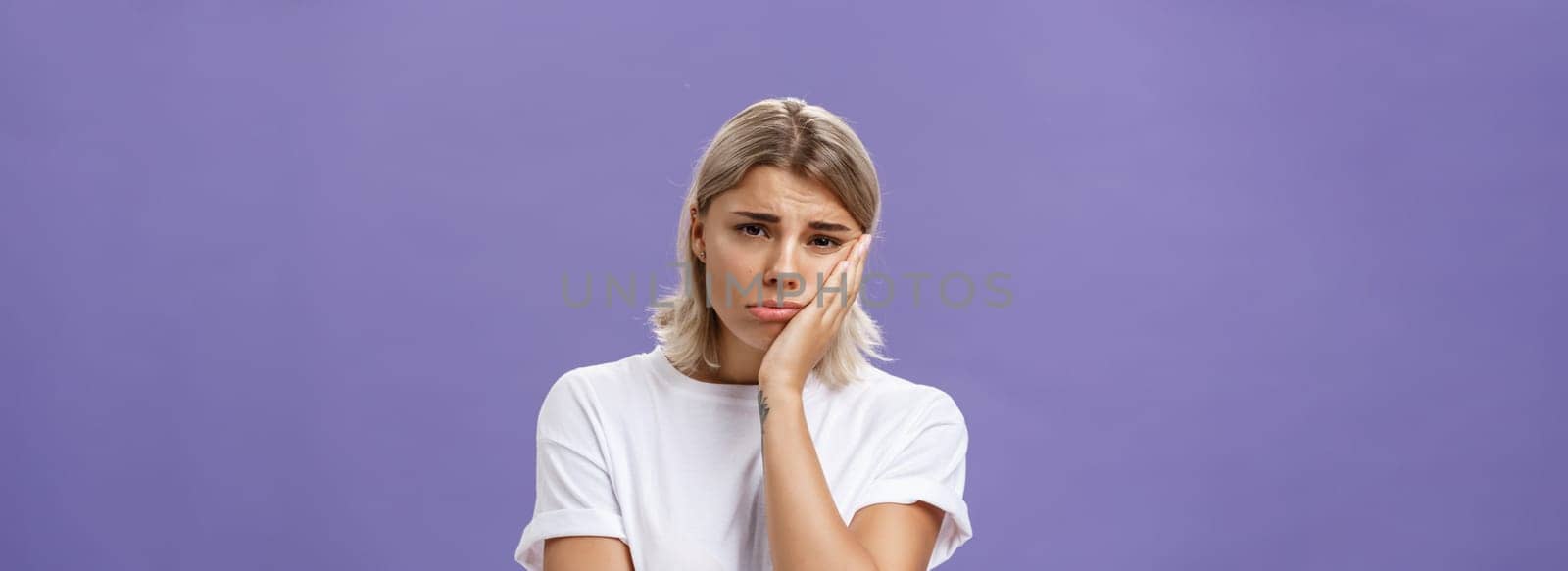 Waist-up shot of unhappy miserable and sad cute blonde female in white casual t-shirt pursing lips leaning face on palm and frowning from disappointement and regret over purple background. Copy space