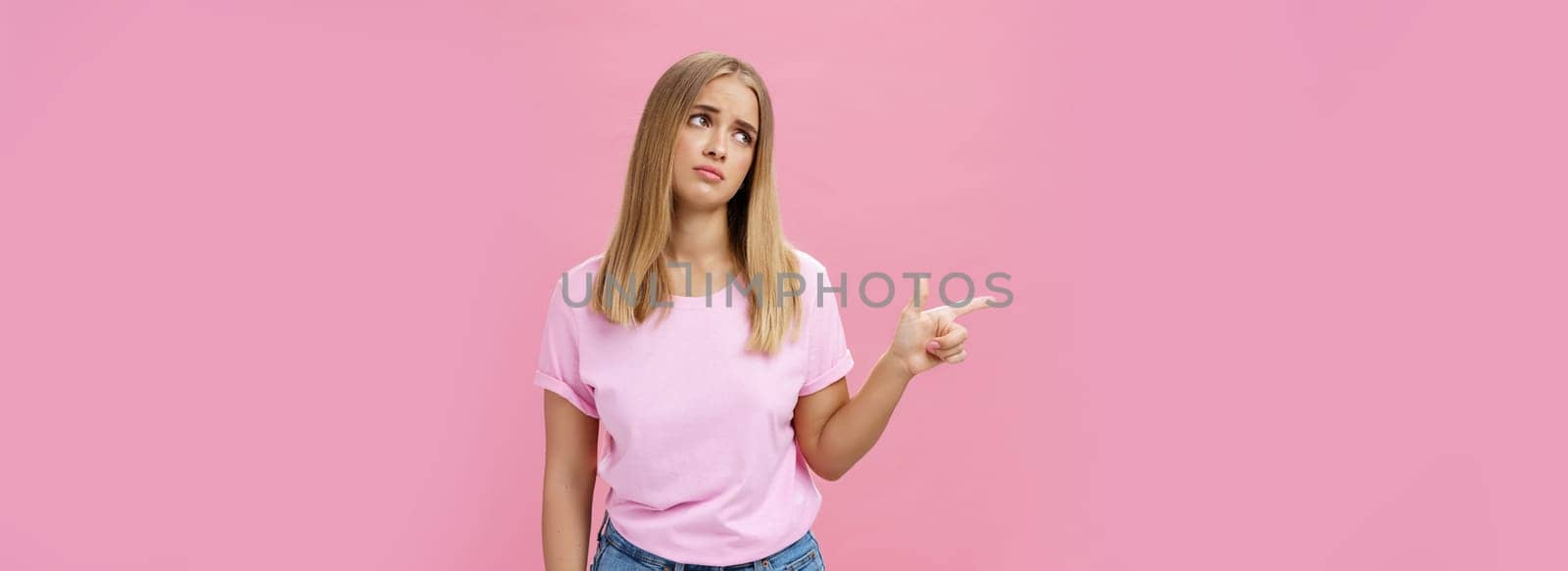 Envy upset cute young european woman with tanned skin and fair hait tilting head lifting eyebrows in sad silly look pointing, gazing left with regret and disappointment posing against pink background.