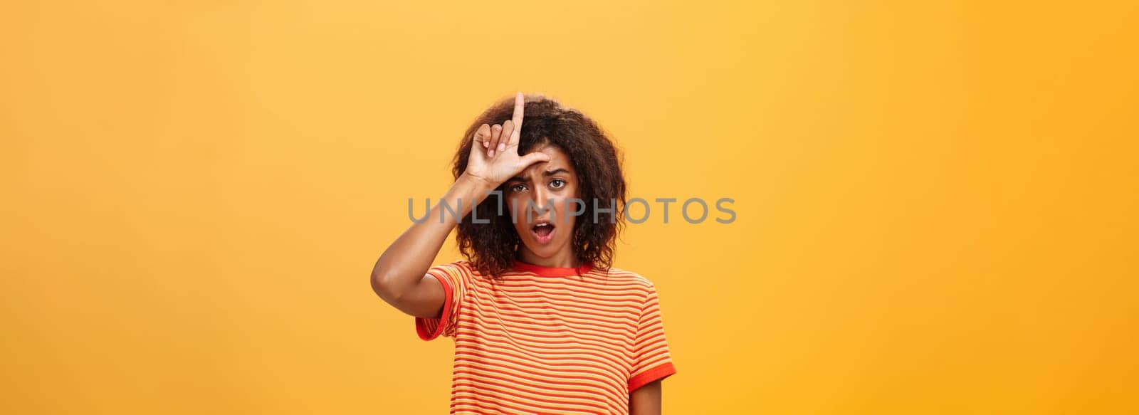 Girl thinks she loser. Portrait of gloomy bothered and displeased african american woman with afro hairstyle showing l word over forehead complaining feeling gloomy and unhappy over orange background by Benzoix