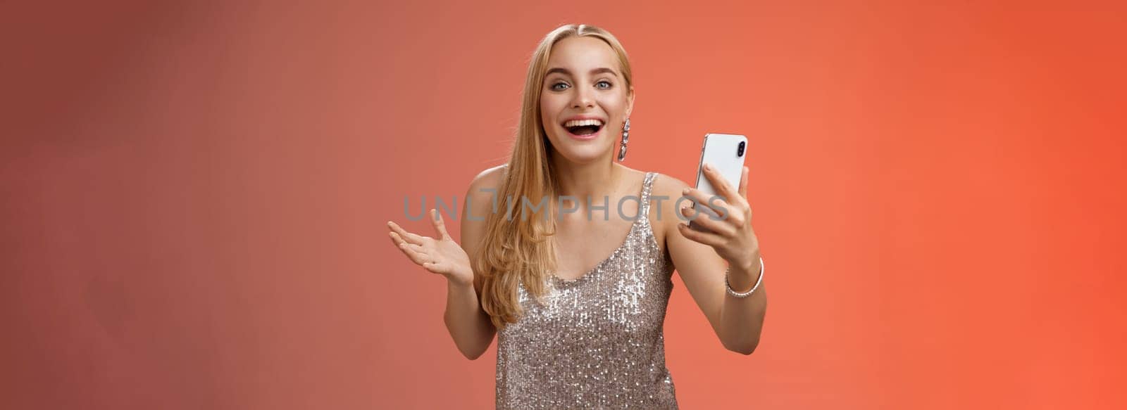 Astonished happy charming blond girl in silver glittering stylish dress holding smartphone amazed liking awesome result edit photo app smiling wondered amused, standing red background by Benzoix