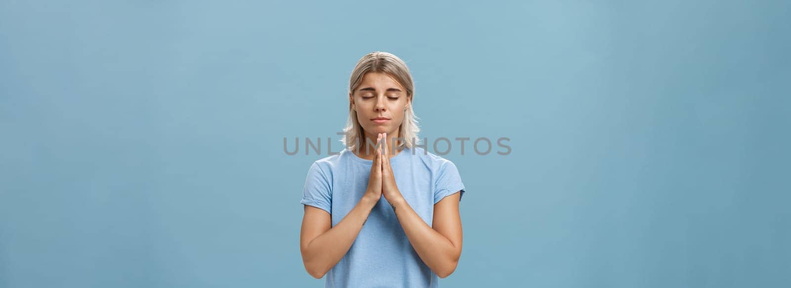 Good-looking kind and faithful european girl with blond hair smiling cute and tender holding hands in pray while making wish hopefully believing god hearing her prayers with closed eyes over blue wall by Benzoix
