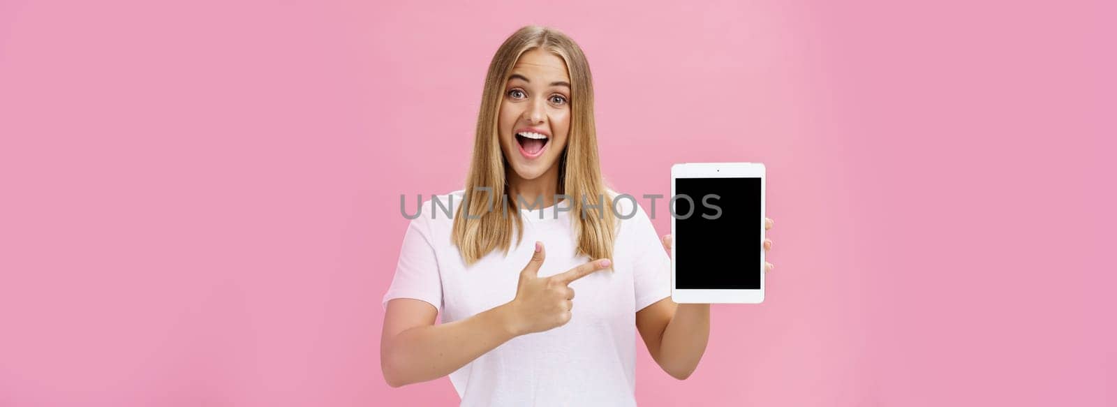 Woman giving recommendation buy digital tablet pointing at device screen with index finger smiling enthusiastic and pleased telling own opinion about gadget perfomance posing delighted over pink wall by Benzoix