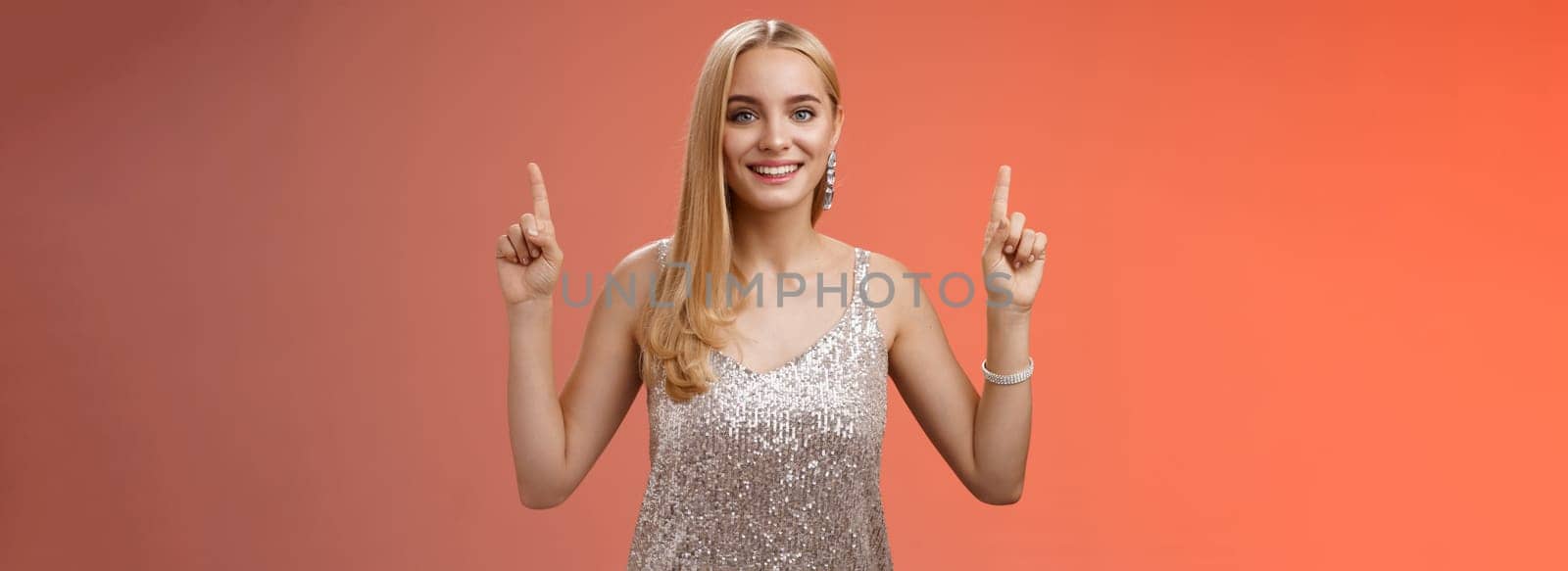 Charming feminine tender blond woman in silver party dress raise hands pointing up smiling delighted recommend awesome cosmetics good product service, standing happily grinning red background.