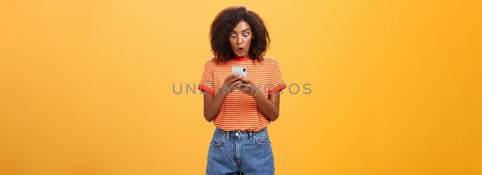 Indoor shot of shocked stunned african american young woman with afro hairstyle staring surprised and excited at smartphone screen holding cellphone reading amazing message over orange wall. Lifestyle.