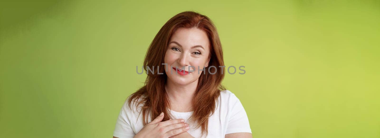 Lovely pleased tender redhead middle-aged ginger woman press hands heart feel touched grateful appreciate sincere heartwarming moment admire thankfully grin look camera glad green background by Benzoix