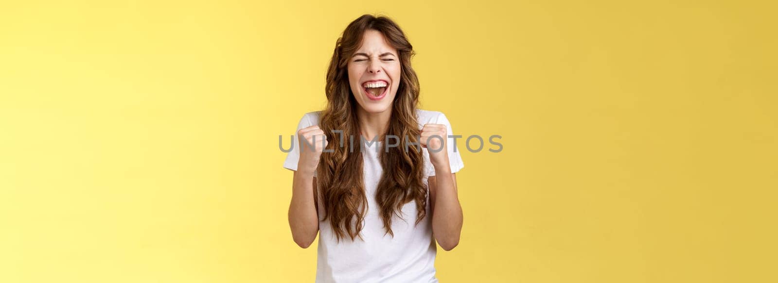 Excited happy triumphing joyful caucasian girl close eyes fist pump celebratory happiness gesture yelling yeah success reach goal achievement dancing victory winning feel relieved yellow background. Lifestyle.