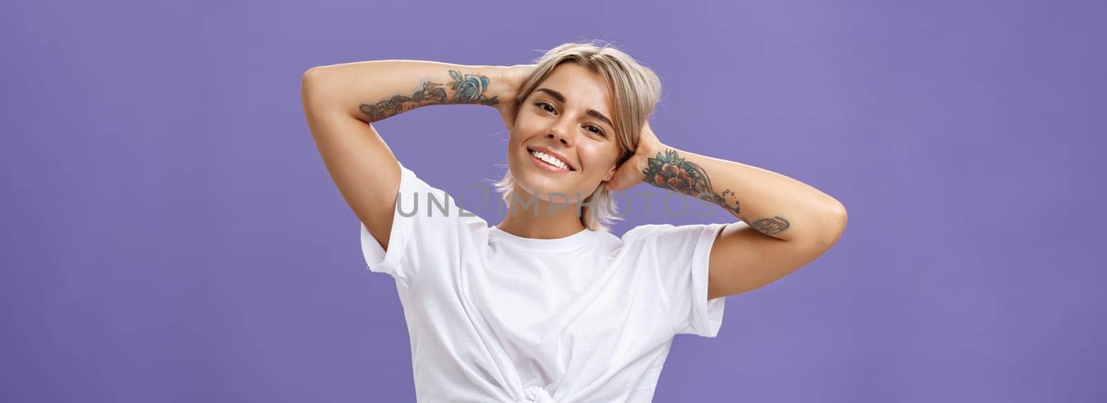 Lifestyle. Close-up shot of good-looking stylish and relaxed blond woman with tattoos on arms holding hands behind head smiling with carefree pleased look enjoying weekends over purple background.