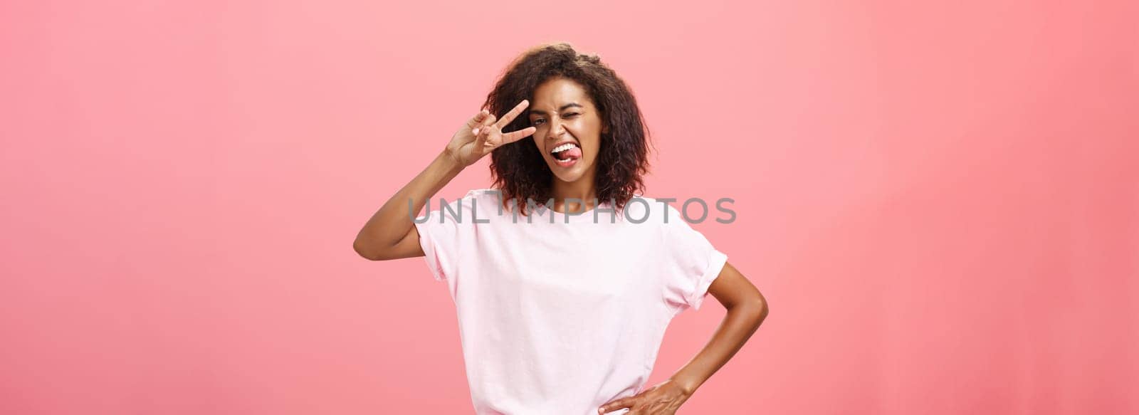 Not afraid express myself. Joyful charismatic african american woman in t-shirt with afro haircut showing tongue playfully and daring making peace sign over eye and winking posing over pink background by Benzoix
