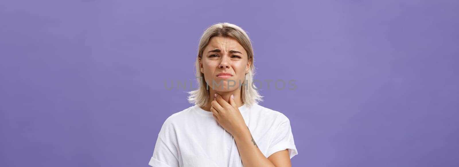 Displeased young stylish caucasian woman with blond hair and tanned skin feeling discomfort in throat having inflammation or feeling cold touching neck and frowning dissatisfied from pain by Benzoix
