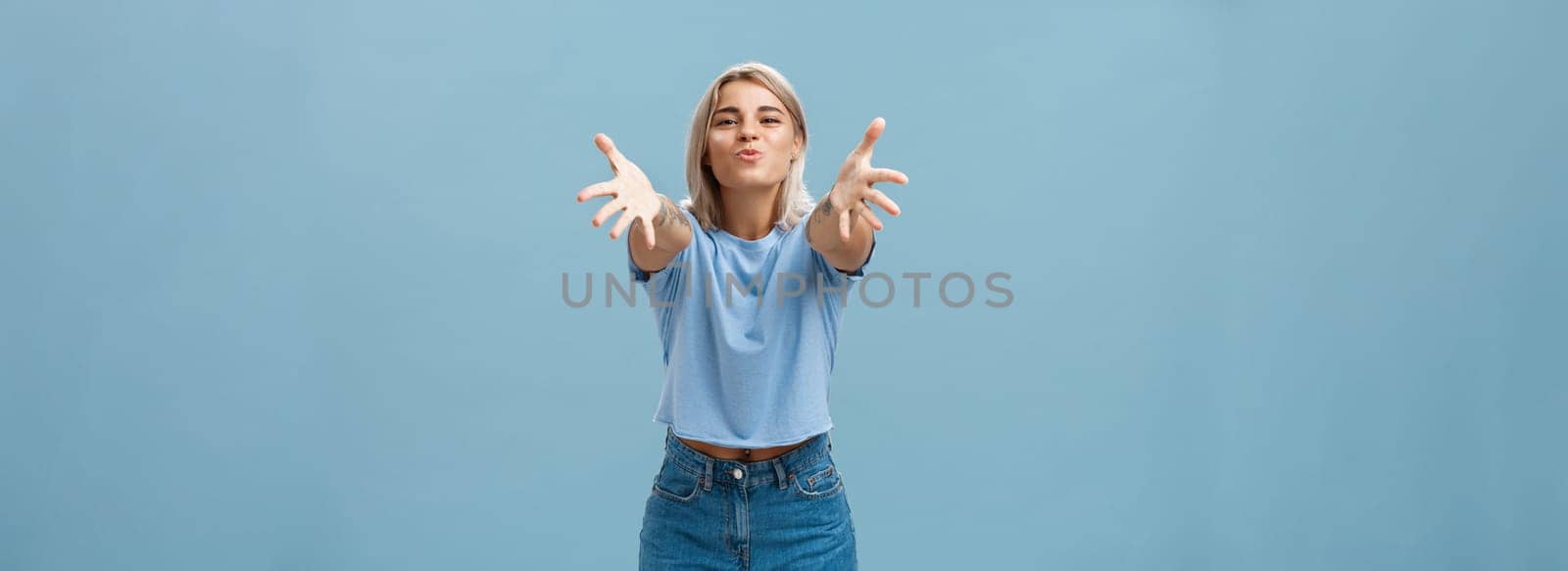 Come to mama. Portrait of friendly joyful and enthusiastic caucasian female student in trendy outfit pulling hands towads camera and folding lips to hug and give passionate kiss smiling over blue wall.