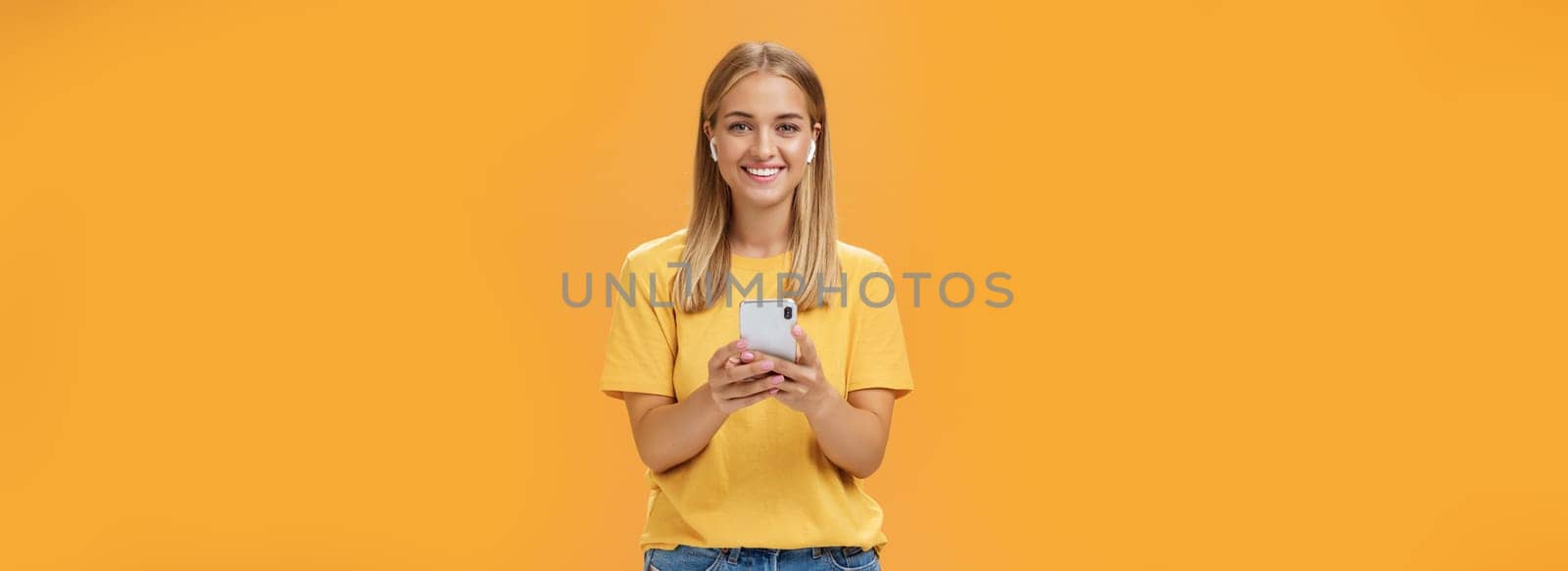 Portrait of friendly charismatic and outgoing good-looking young female in yellow t-shirt holding smartphone wearing wireless earphones enjoying listening music on way to fitness gym. Technology and lifestyle concept