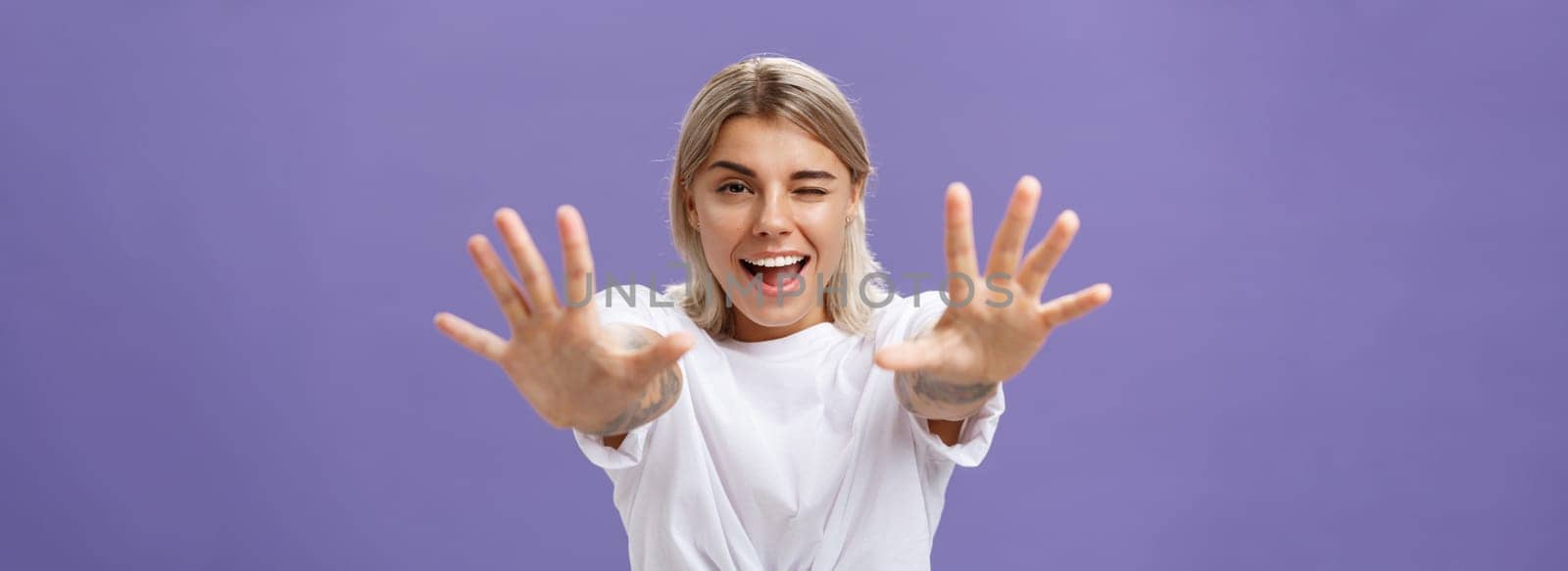 Reaching to you. Portrait of flirty good-looking and confident stylish woman with tattoos on arms winking sticking out tongue playfully and smiling pulling hands towards camera over purple background. Emotions and body language concept
