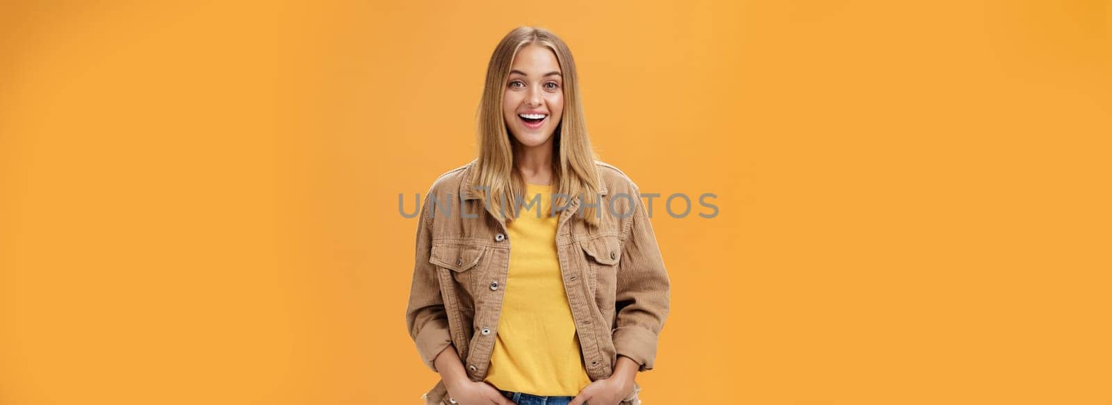 Charismatic tanned woman in corduroy jacket and yellow t-shirt ready for chilly autumn walk with friends smiling joyfully gazing entertained at camera holding hand in pockets casually over orange wall. Lifestyle.