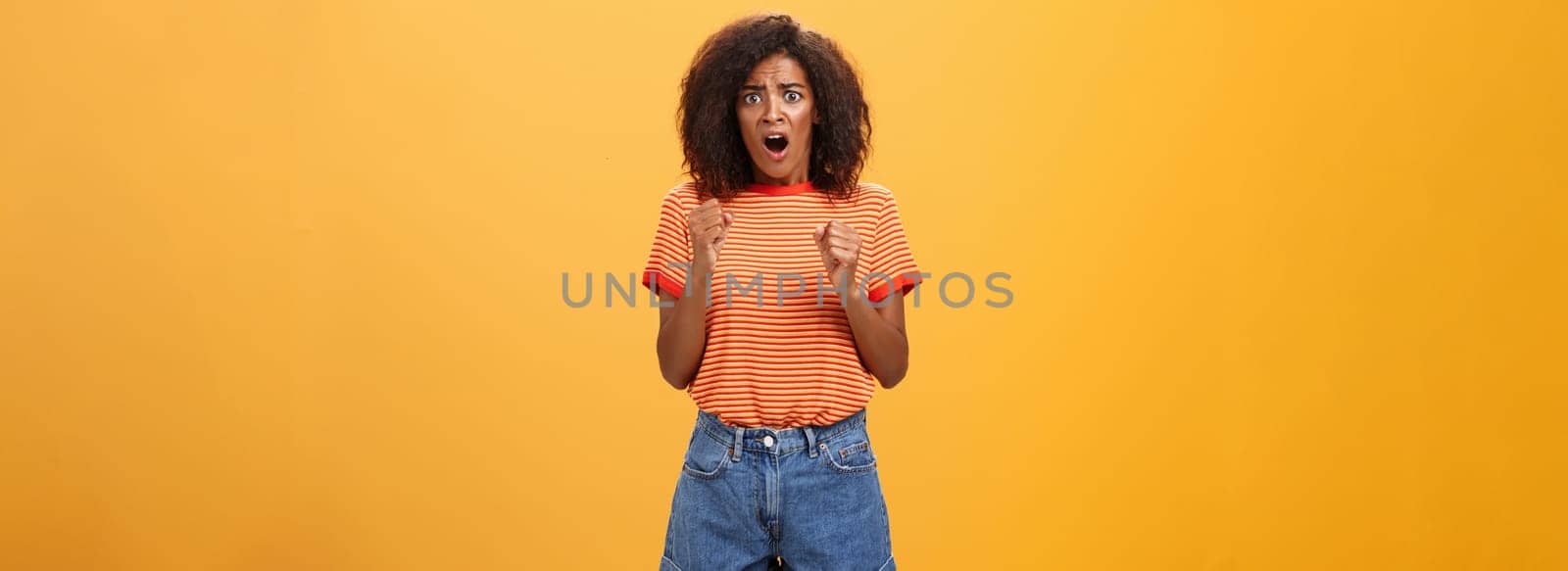 Woman about to panic hearing shocking worried news. Portrait of intense nervous and anxious dark-skinned adult girl with afro hairstyle clenching fists opening mouth looking concerned over orange wall by Benzoix