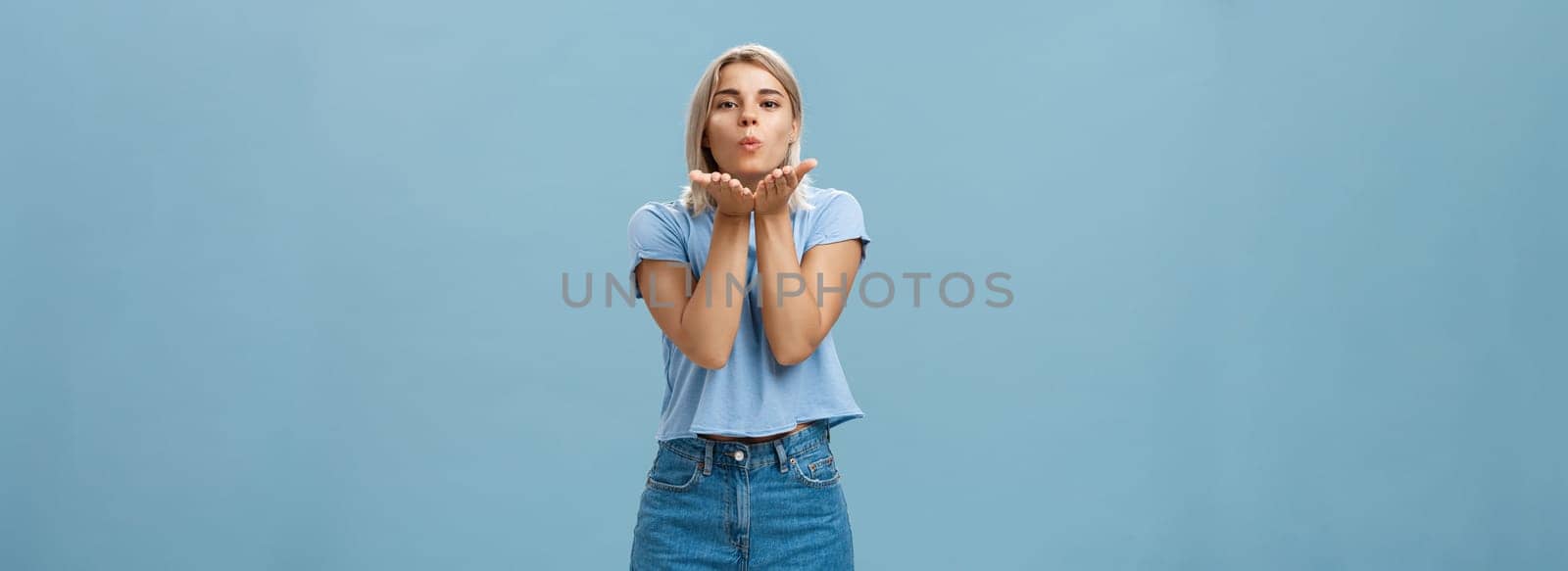 Lifestyle. Studio shot of attractive and romantic european blonde girlfriend in denim shorts folding lips sending wind kiss with palms near mouth gazing tender and cute at camera over blue background.