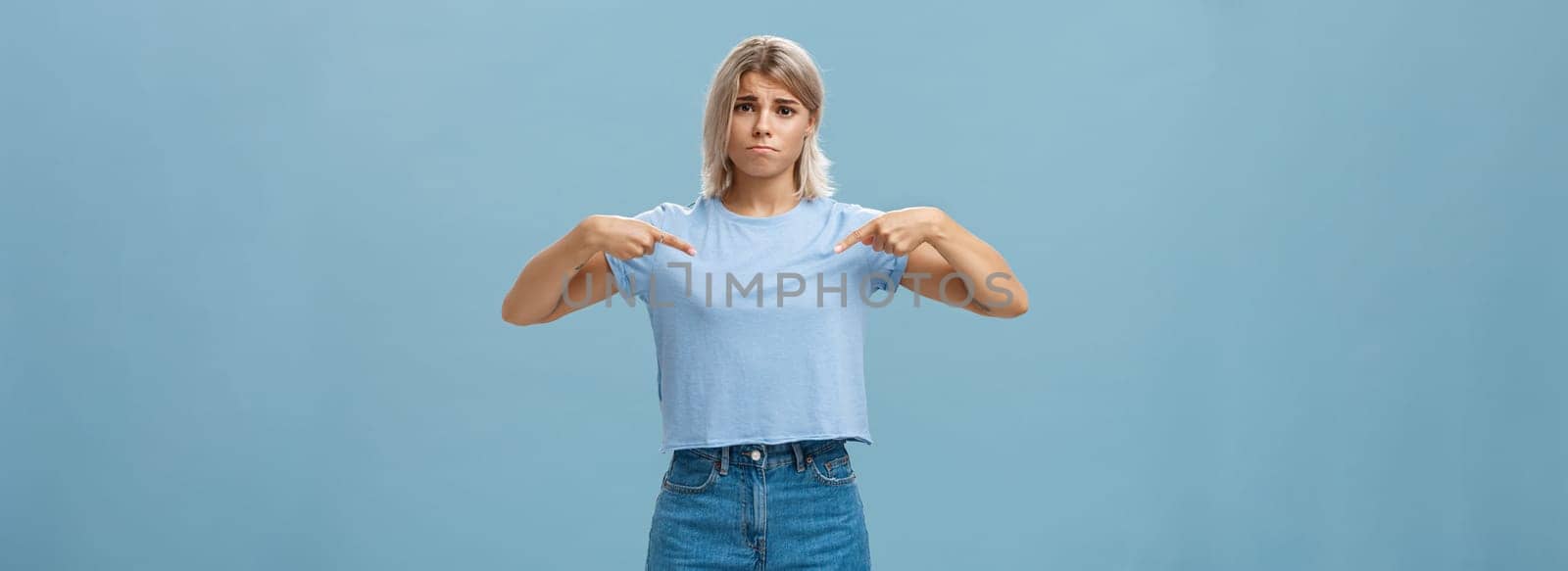 Sad cute blonde young female complaining being displeased with own body, pointing at breast frowning making gloomy smile pulling lips down from disappointment standing over blue background by Benzoix