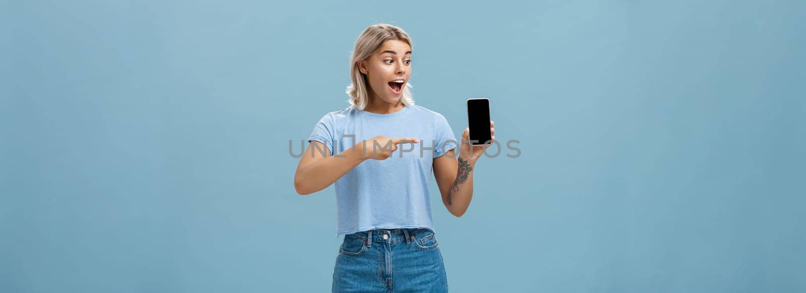 Look at this awesome smartphone. Impressed enthusiastic good-looking female blonde with cool tattoo on arm dropping jaw from thrill and excitement pointing at device screen over blue background.