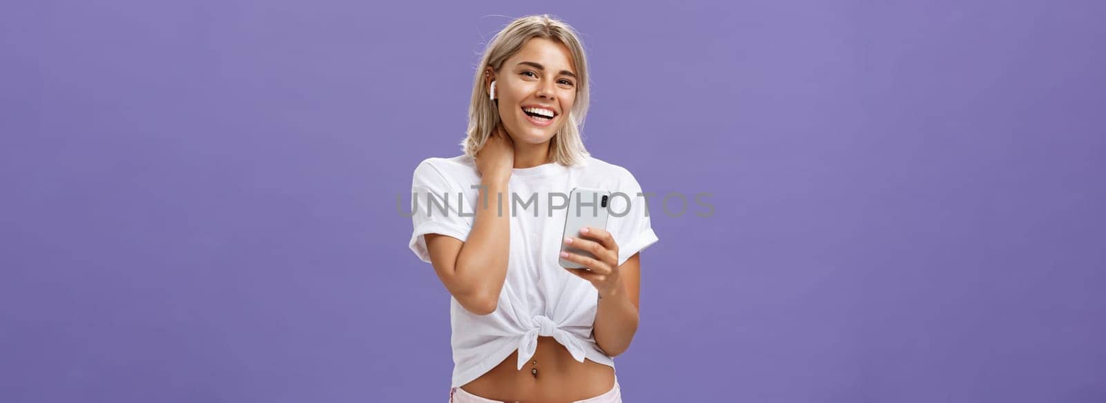 Shy attractive and stylish sociable woman in white t-shirt touching neck timid and awkward smiling broadly wearing wireless earphones while listening songs with smartphone over purple wall. Technology concept