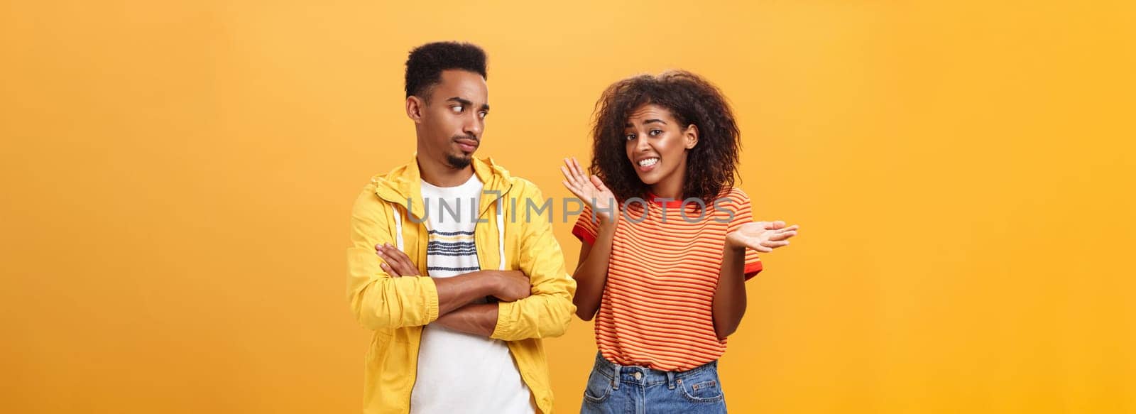 Guy thinks his friend weirdo making dumb thinks looking at cute girl with suspicious look crossing arms on chest raising eyebrow questioned while girlfriend saying sorry shrugging over orange wall by Benzoix