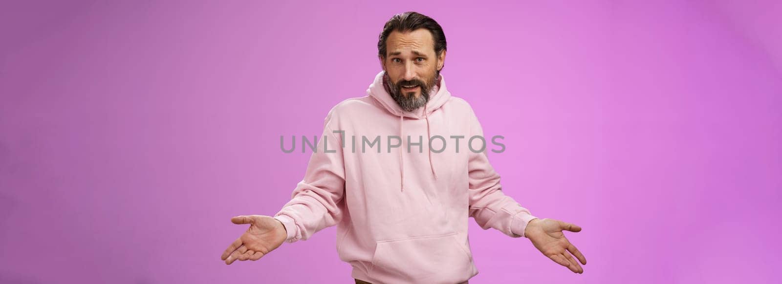 Bothered confused mature adult bearded man grey hair in pink hoodie arguing look pissed offended shrugging spread hands sideways dismay look clueless camera asking why, purple background.