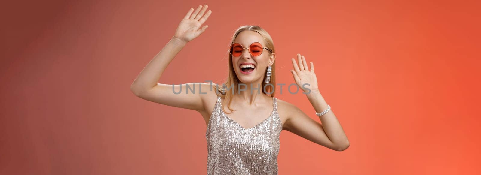 Happy amused carefree blond woman go wild dance-floor dancing having fun yelling yeah closed eyes waving hands moving rhythm music joyfully party in silver stylish dress sunglasses, red background.