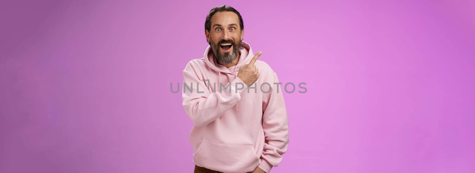 Speechless excited impressed handsome bearded man grey hair widen eyes surprised astonished wow drop jaw gasping thrilled pointing upper left corner see famous star, purple background.