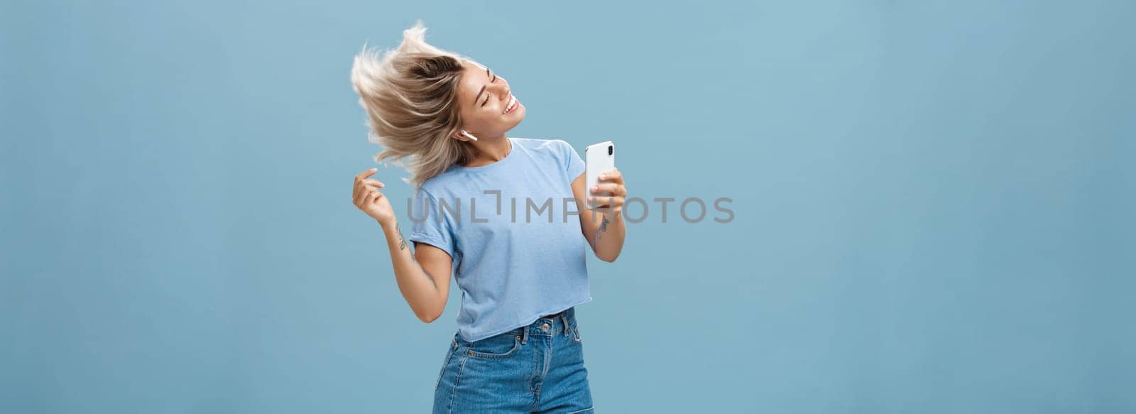 Girl enjoying cool bits in brand new wireless earphones advertising earbuds in own blog recodring video via smartphone dancing from joy and delight smiling listening music over blue background by Benzoix