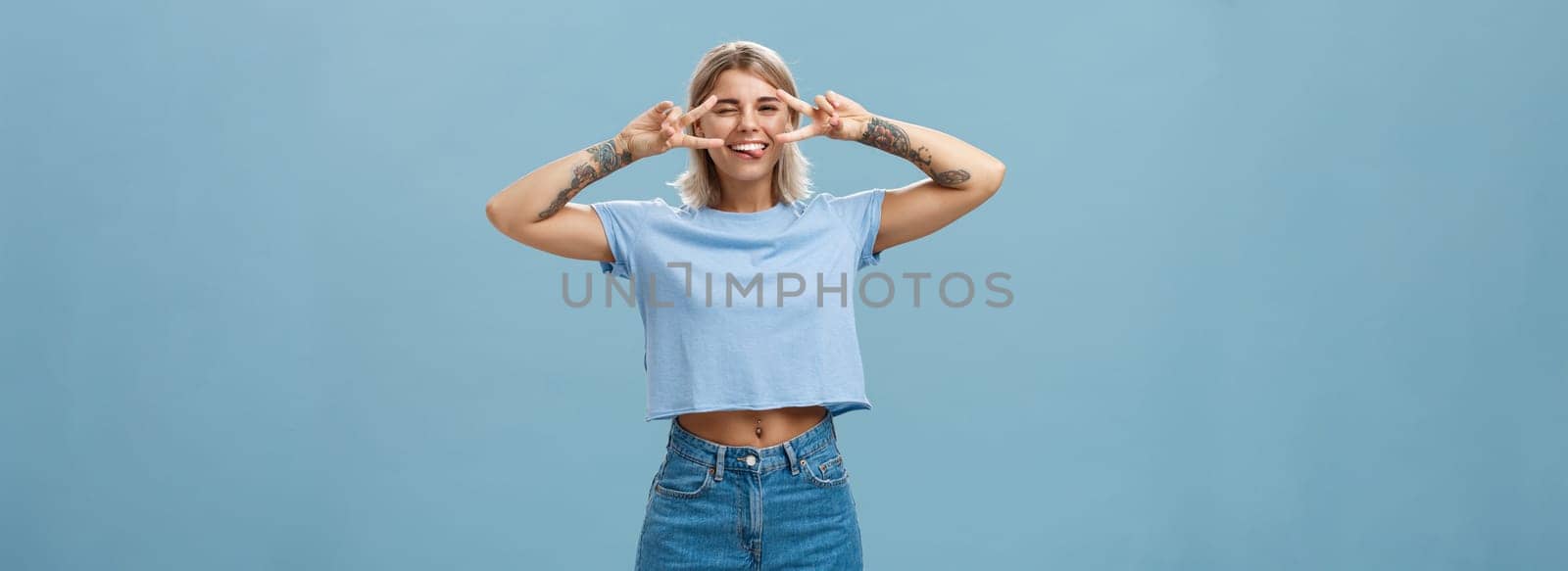 Enjoy life and dream. Portrait of joyful good-looking carefree european female student with blond short haircut winking sticking out tongue entertained and showing peace signs over eyes near blue wall by Benzoix