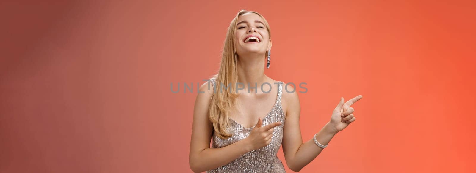 Funny carefree blond european woman in silver glittering evening dress raise head up close eyes laughing happily pointing left see hilarious person joking fool around, standing red background.
