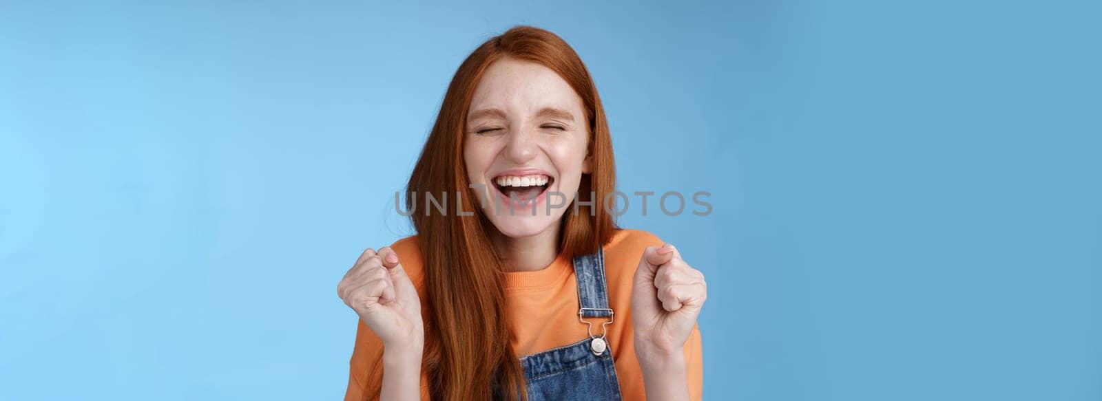 Sincere happy rejocing ginger girl close eyes smiling broadly say yes waving clenched fists joyfully celebrate enterting university dream come true winning prize triumphing cheerfully blue background by Benzoix