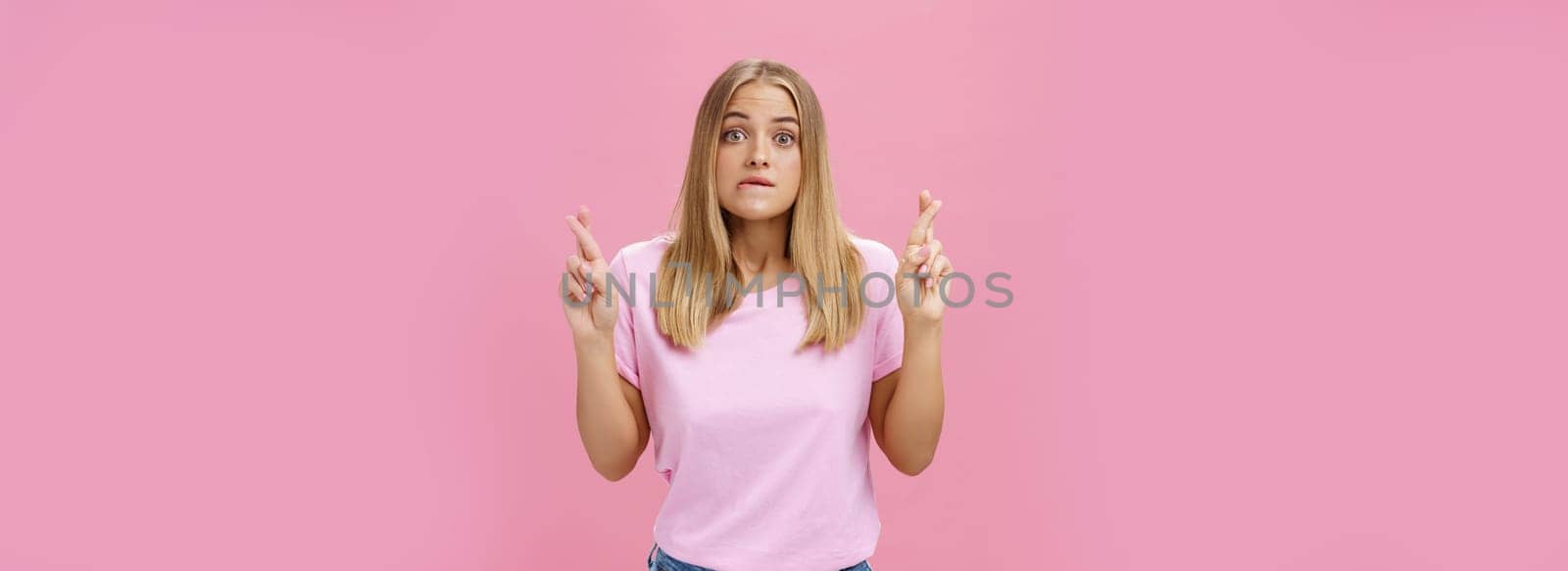 Woman hopefully looking at chart praying with crossed fingers to pass exam biting lip popping eyes, standing intense and worried against pink background having hope to win or receive scholarship by Benzoix