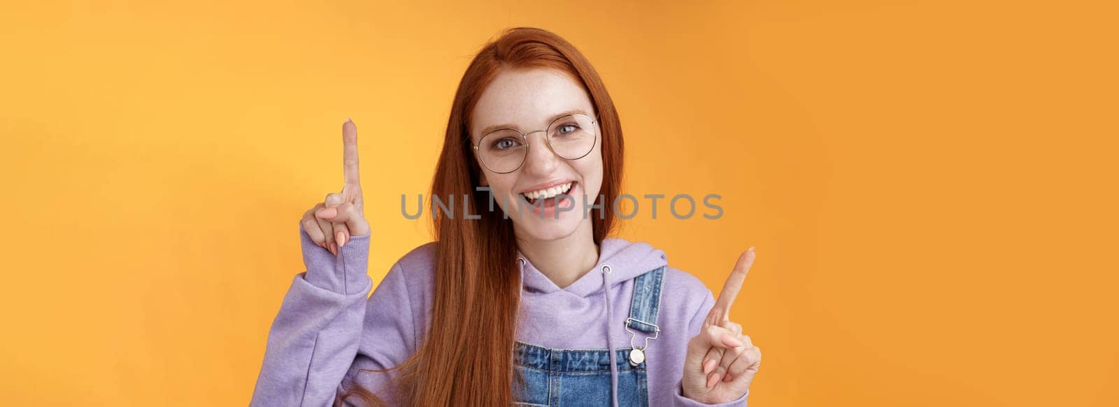 Lively enthusiastic millennial redhead girl coworker having fun celebrating small break joyfully dancing pointing up index fingers singing smiling white teeth promoting product, orange background.