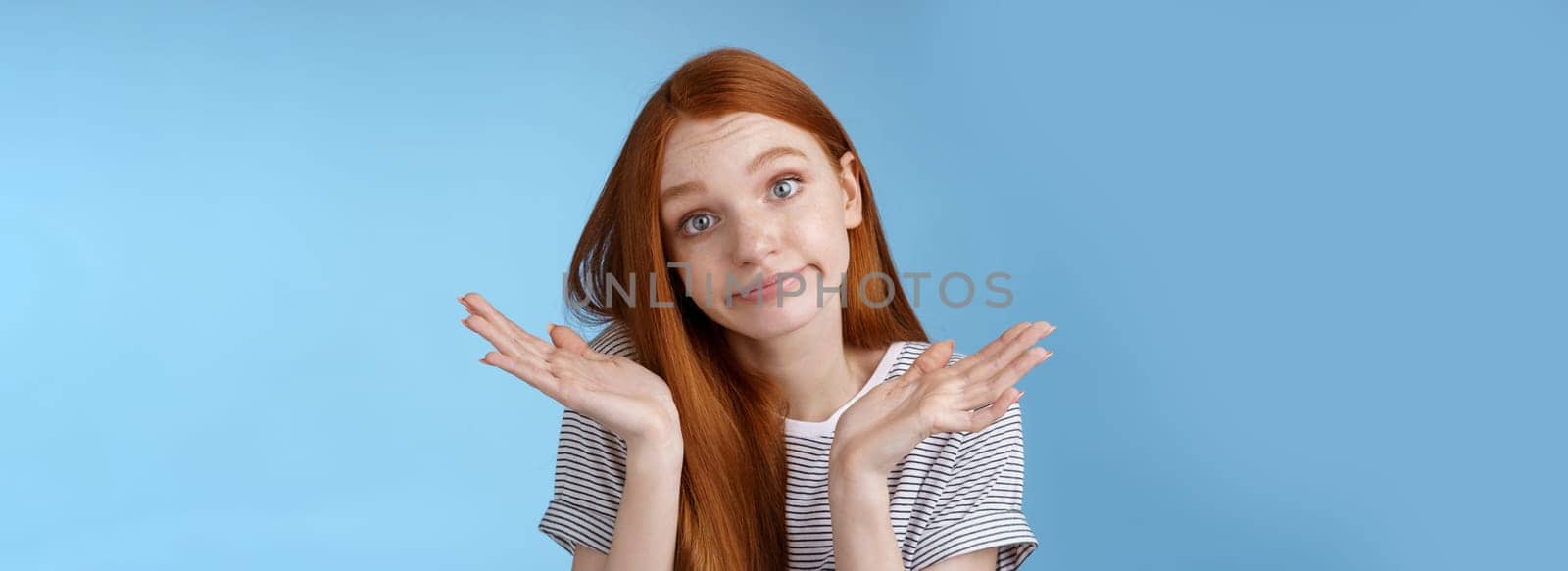 Unsure clueless cute silly redhead female part-time worker shrugging careless raise hands unaware smirking tilting head lift eyebrows confused cannot answer no idea standing blue background by Benzoix