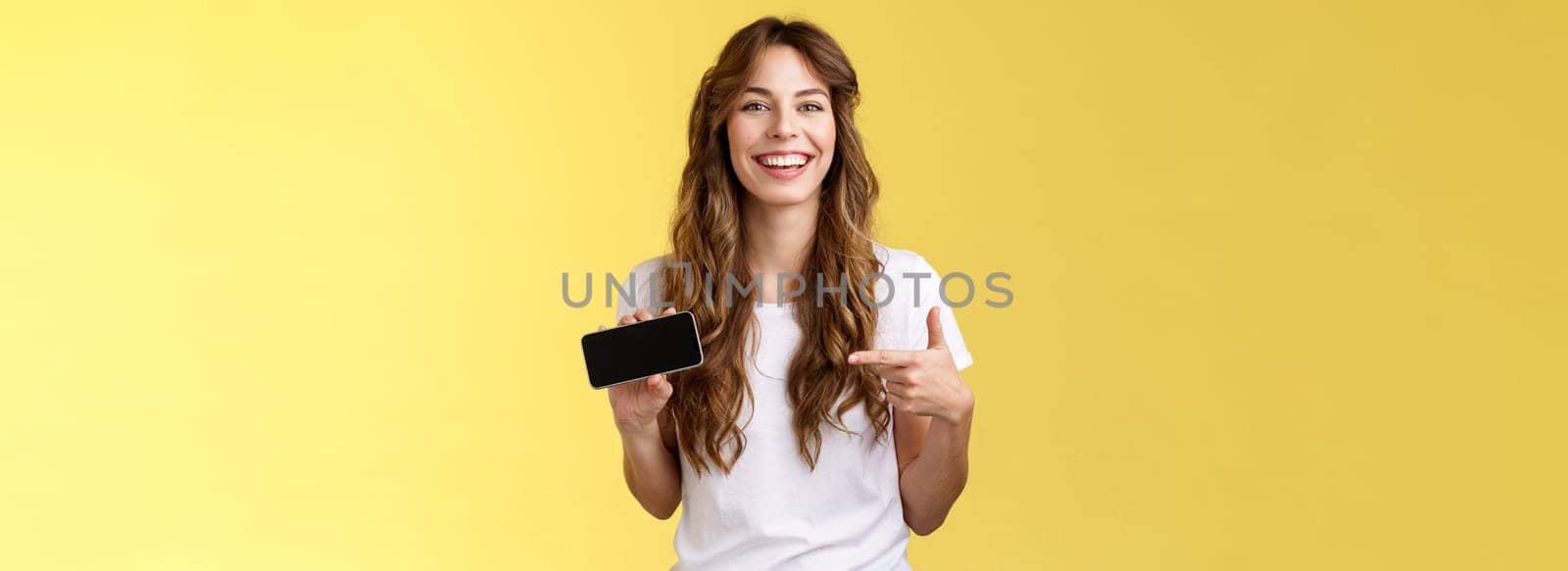 Carefree charismatic cute curly-haired female gamer likes playing smartphone games showing own game score hold mobile phone horizontal pointing display laughing pleased amused. Copy space