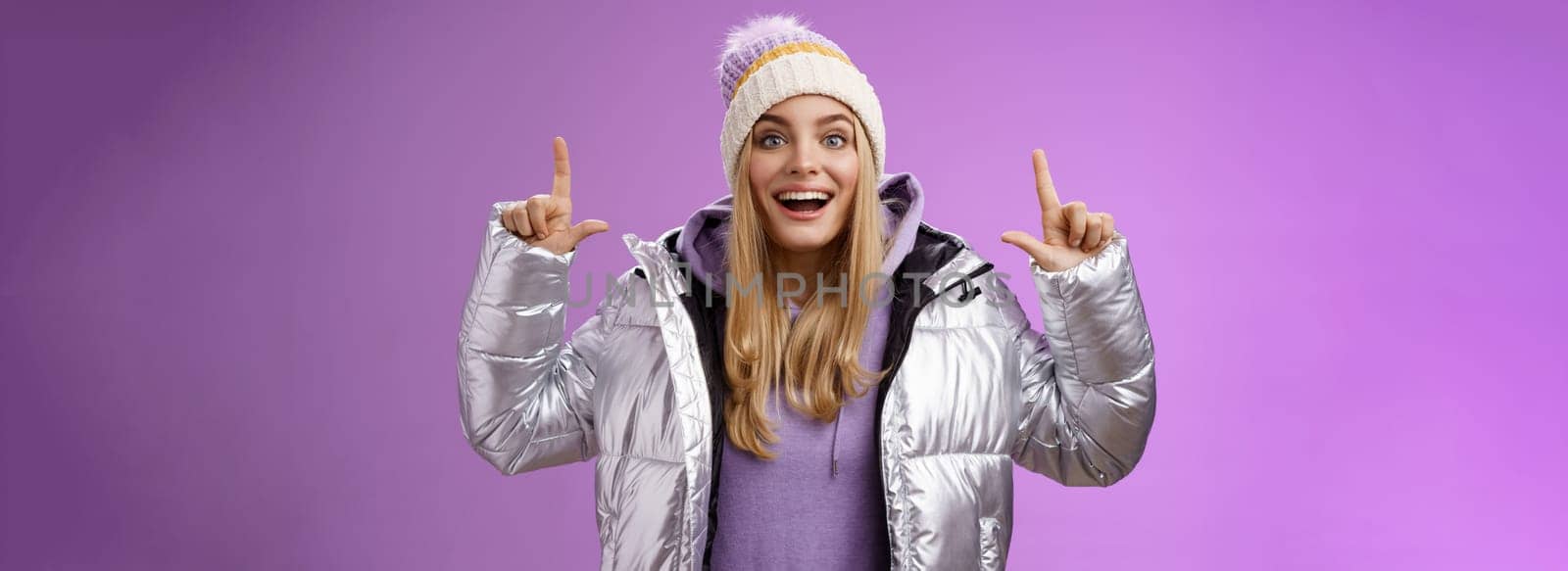 Lifestyle. Excited carefree cheerful fair-haired european girl in silver jacket winter hat raising hands pointing up have excellent idea smiling broadly speaking passionately standing purple background.