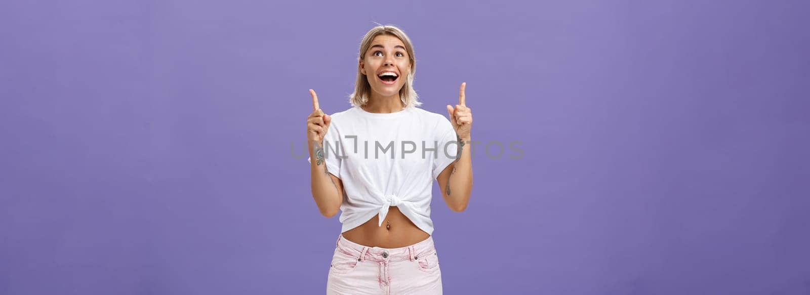 Lifestyle. Woman seeing miracle being impressed and delighted like child staring and pointing up with broad thrilled smile standing over purple background in summer trendy outfit being joyful and excited.