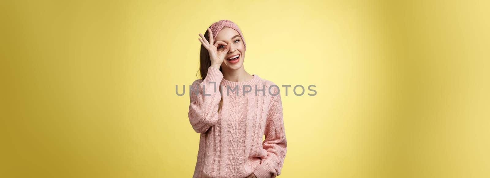 Cheerful girl saying ok. Attractive stylish shoolgirl in knitted sweater relaxed feeling upbeat, sharing happiness and joy, showing okay sign on eye smiling optimistic, entertained over yellow wall.