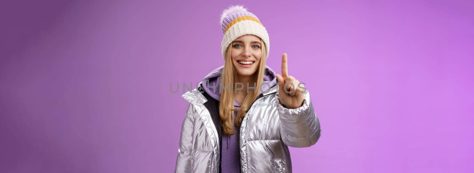 Excited cheerful fair-haired european girl in winter hat silver shiny jacket show number one index fingers give suggestion advice smiling broadly talking happily, purple background. Copy space
