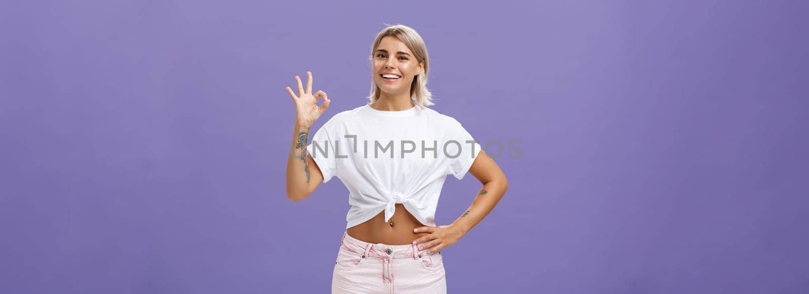 Excellent I like it. Portrait of satisfied good-looking happy girl with fair hair in white t-shirt and shorts showing ok or perfect gesture and smiling broadly holding hand on waist over purple wall by Benzoix