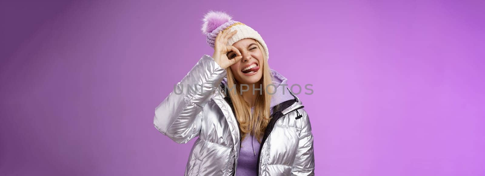Friendly joyful caucasian blond girl in hat warm stylish shiny silver jacket tilt head happily show tongue smiling broadly enjoying awesome ski resort vacation travelling winter, purple background by Benzoix