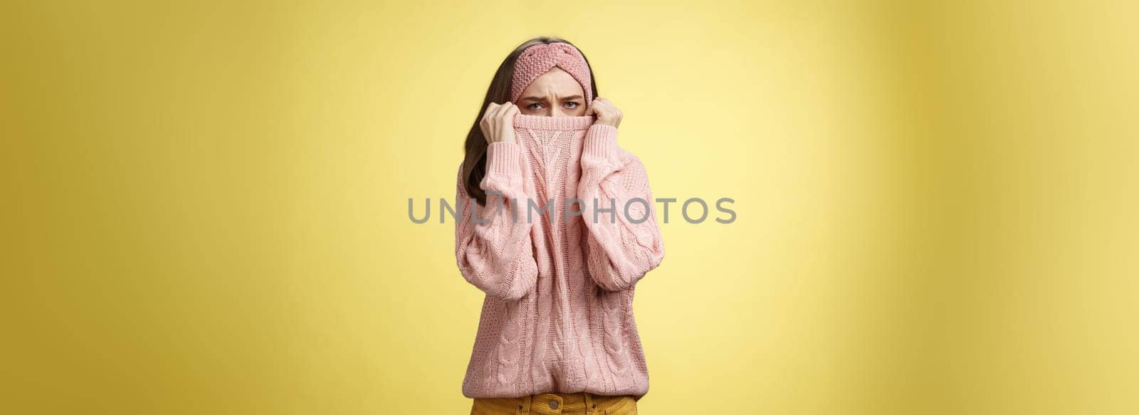 Sad silly, gloomy cute young girl hiding face in sweater collar pulling clothes on nose frowning displeased, unhappy, looking indignant and reluctant standing disappointed over yellow background.