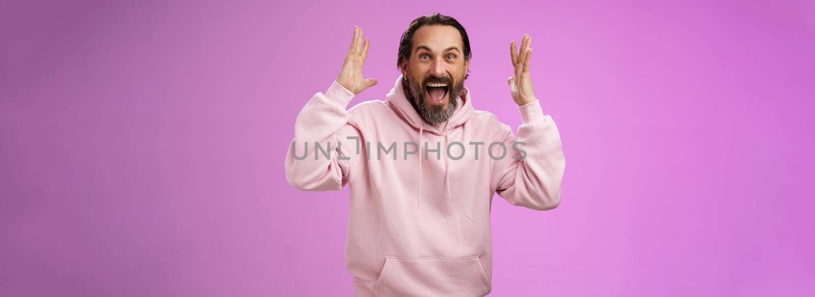 Happy excited lucky adult bearded man celebrating excellent news raising hands gesturing thrilled smiling broaldy triumphing victory win, yelling gladly achieve goal, standing purple background.