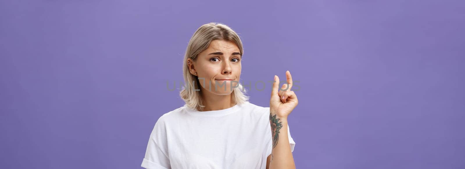 Girl having tiny problem. Concerned attractive blonde girl with tattoo on arm pursing lips in troubled look shaping small or little object, dissatisfied with regret in eyes over purple wall. Copy space