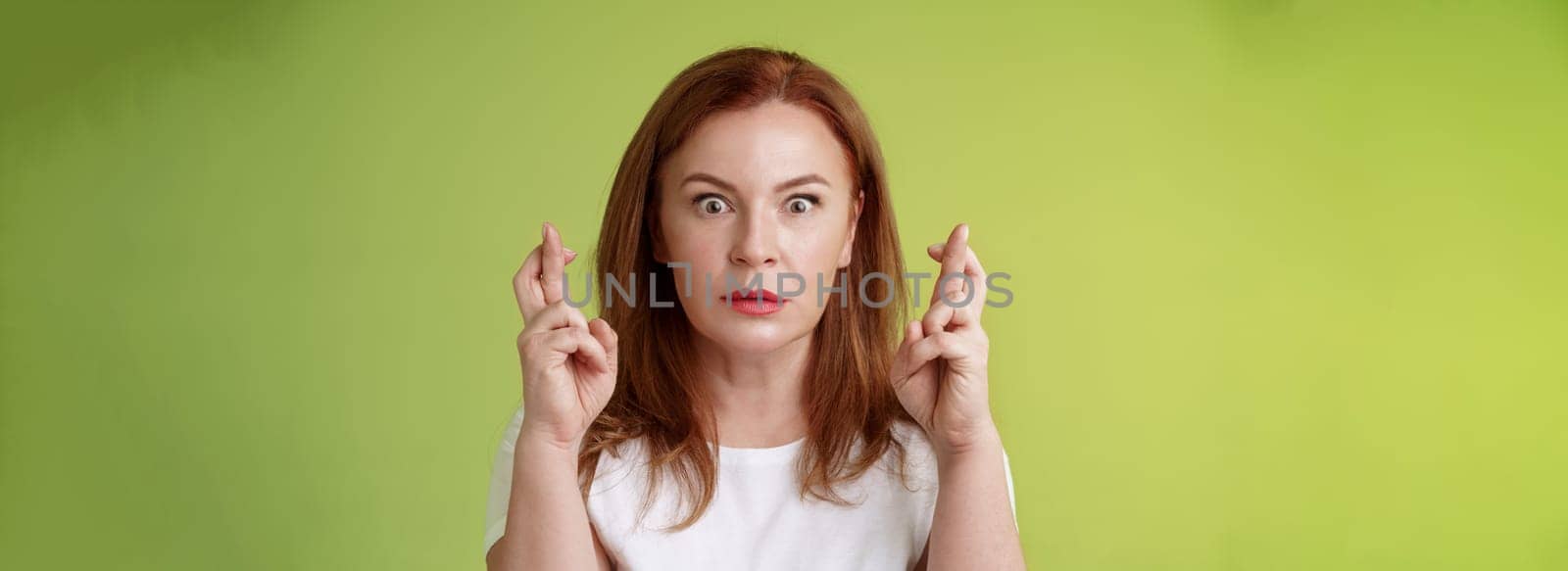 Stunned intense worried middle-aged redhead woman stare nervously awaiting important result implore dream come true receive positive reply standing stupor green background make wish good luck by Benzoix