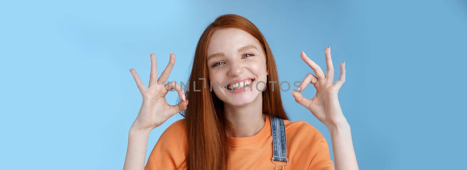 Wellbeing, happiness, perfection concept. Attractive friendly smiling joyful redhead female student show excellent okay ok gestures grinning approval agree awesome concept, satisfied blue background by Benzoix