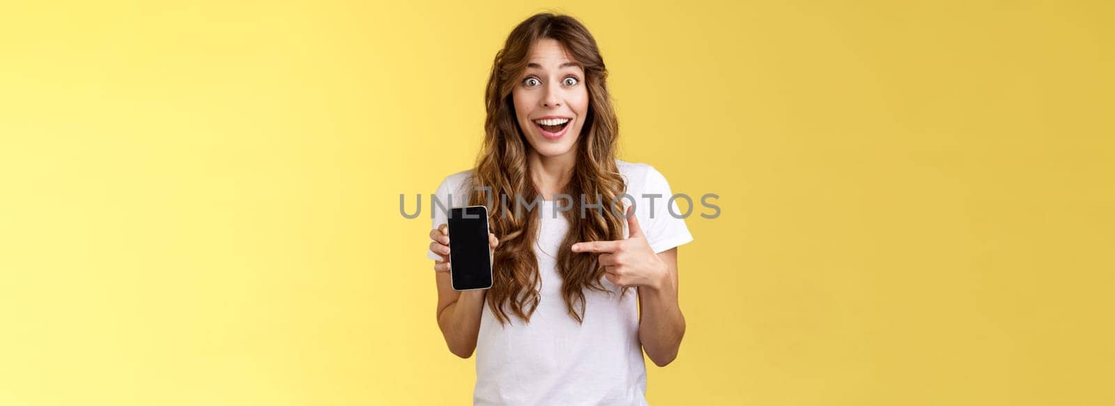 Surprised happy lucky girl winning online internet lottery smiling broadly hold smartphone pointing blank mobile phone screen showing display grinning excited enthusiastic stand yellow background by Benzoix
