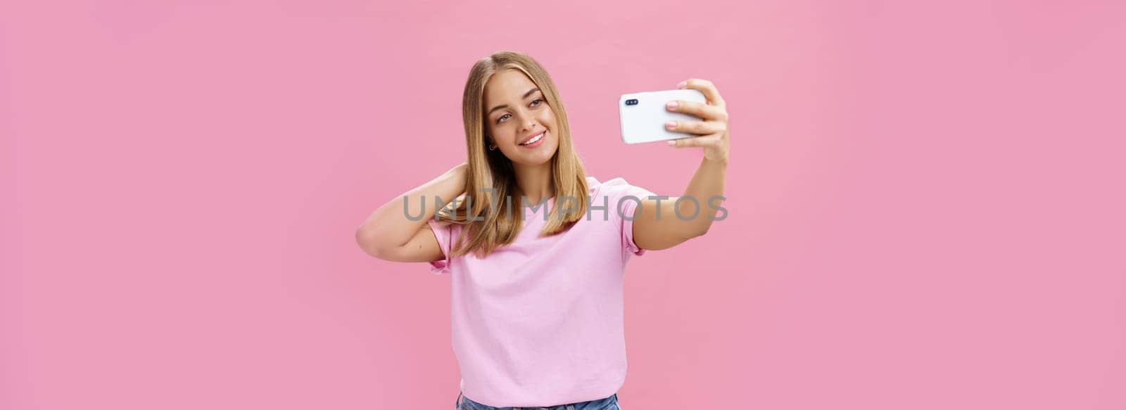 Female beauty blogger taking selfie post new look online. Portrait of charming tanned young woman in t-shirt touching hair gently pulling hand with smartphone near face taking photo over pink wall. Technology concept