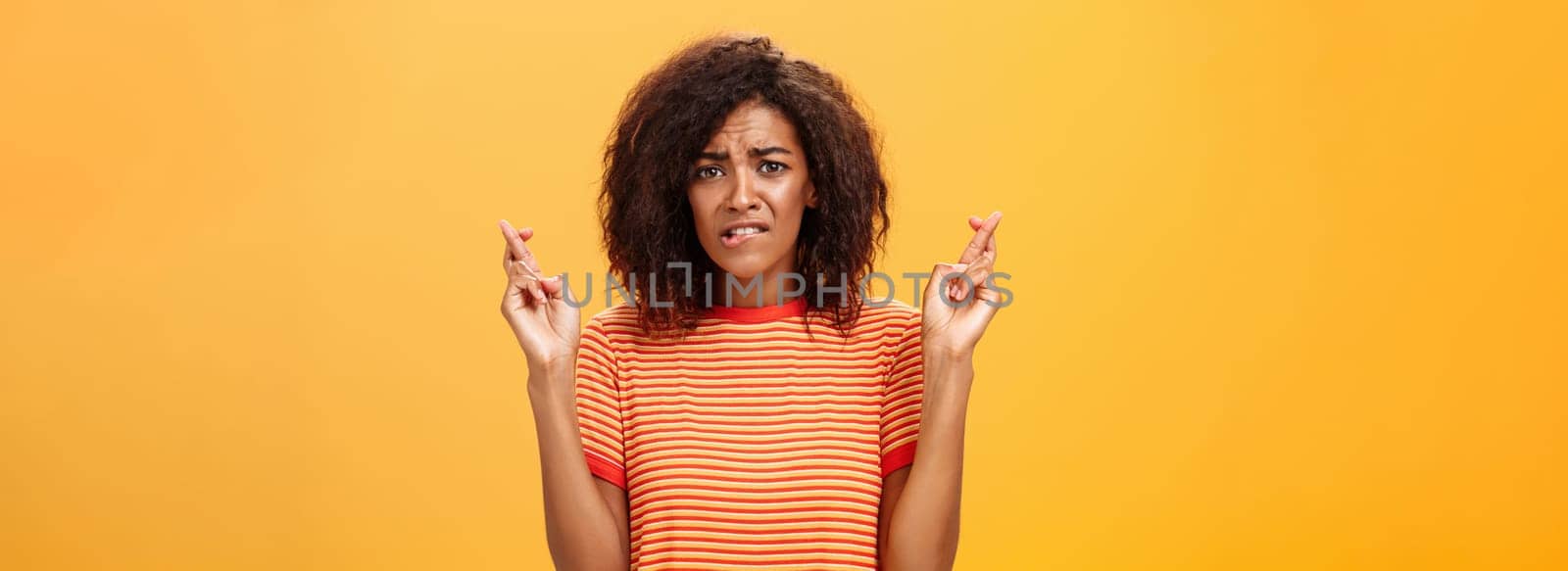 Waist-up shot of concerned troubled nervous african american woman with curly hairstyle biting lower lip anxiously frowning crossing fingers for good luck hopefully praying for dream come true. Lifestyle.
